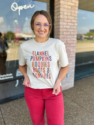 Woman smiling in a cream graphic tee reading "Flannel Pumpkins Hoodies Boots & Bonfires" outside boutique.