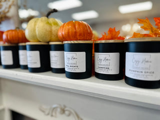 Fall-themed soy candles in black jars with wooden lids displayed on white shelf with pumpkins and autumn leaves