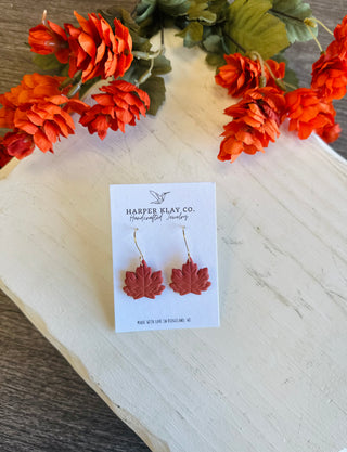 Harper Klay Co. handcrafted clay maple leaf dangle earrings displayed on white card with autumn floral backdrop.