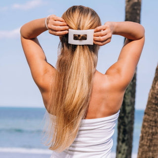Woman styling long blonde hair with white rectangular hair clip at beach