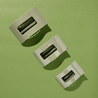 Three sage green rectangular hair clips with clear windows arranged on a green textured background with shadows.