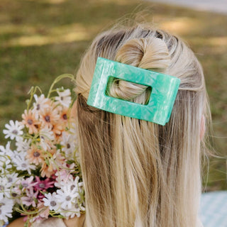 Woman wearing green marbled rectangular hair clip in blonde hair, holding bouquet of white and pink flowers outdoors.