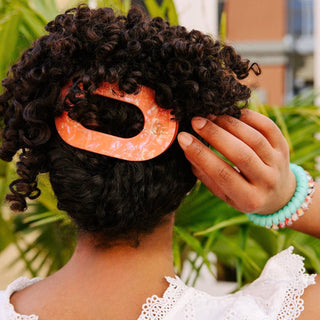 Woman wearing orange flat round hair clip in curly dark hair, hand touching hair accessory.