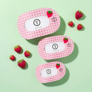 Three pink gingham hair clips with strawberry designs arranged with fresh strawberries on green background.