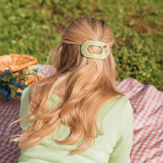 Woman with long wavy hair wearing a gingham pattern flat round hair clip while sitting on picnic blanket outdoors.