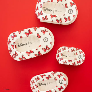 Four oval-shaped Disney TELETIES hair clips with red Minnie Mouse bow pattern on cream background displayed on red surface.