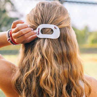 Woman styling blonde wavy hair with white flat round hair clip featuring team logo outdoors