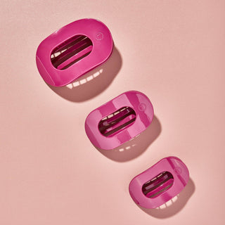 Three pink oval-shaped hair clips with slotted openings arranged on a pink textured surface.
