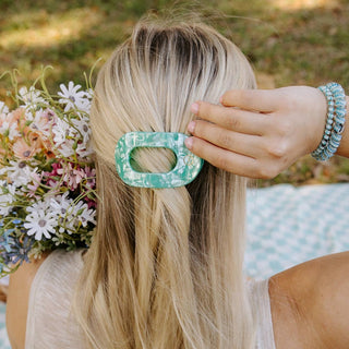 Woman wearing blonde hair with green patterned flat round hair clip, holding flowers outdoors.
