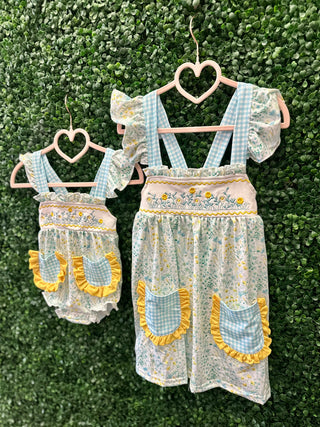 Two matching floral garden dresses with blue gingham straps and yellow ruffled pockets hanging on heart-shaped hangers.