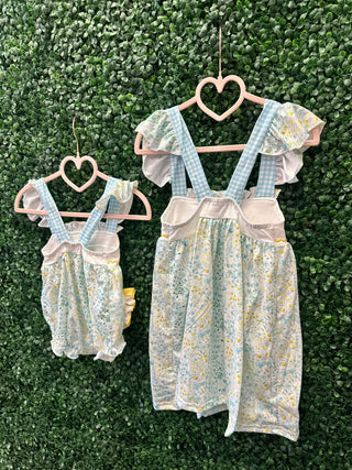Two matching floral garden dresses with blue gingham straps hanging on heart-shaped hangers against greenery backdrop.