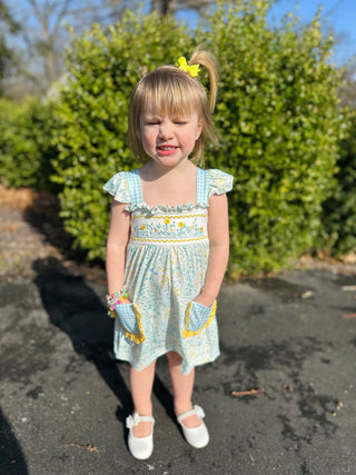Young girl wearing light blue floral dress with yellow trim and pockets, standing outdoors.
