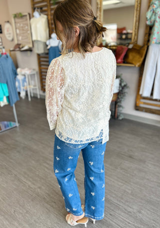 Woman wearing white floral lace tie-front blouse with sheer sleeves and embroidered denim jeans in boutique setting.