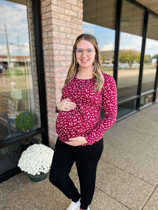 Pregnant woman wearing floral long sleeve maternity nursing top, standing outside brick building storefront.