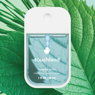 Touchland Power Mist hydrating hand sanitizer spray bottle displayed against lush tropical green leaves background.