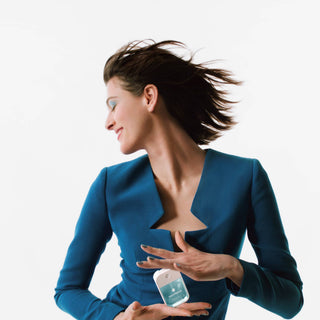 Woman in teal blazer applying Touchland Power Mist hand sanitizer with hair flowing dynamically.