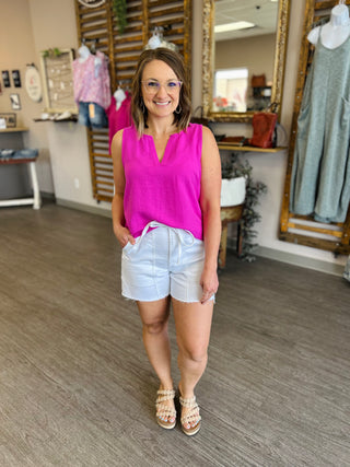 Woman wearing bright pink sleeveless split neck top with white shorts in clothing boutique setting.