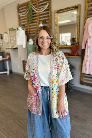 Woman wearing floral patchwork kimono over white top and denim skirt in boutique setting