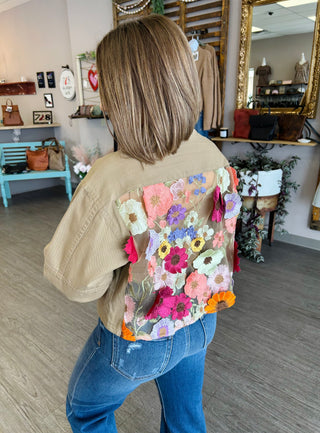 Woman wearing beige denim jacket with colorful embroidered floral design on back in boutique setting.