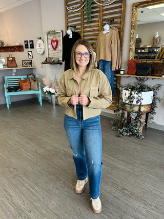 Woman wearing tan denim jacket and jeans standing in boutique clothing store with merchandise displays.