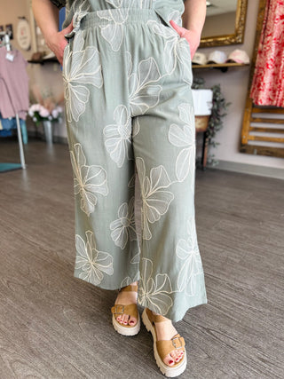 Woman wearing sage green wide-leg pants with white floral pattern and brown platform sandals in boutique setting.