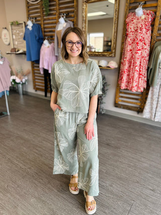 Woman wearing sage green floral print matching top and wide-leg pants set in boutique setting.