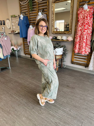 Woman wearing sage green floral print top and matching pants in boutique clothing store setting.