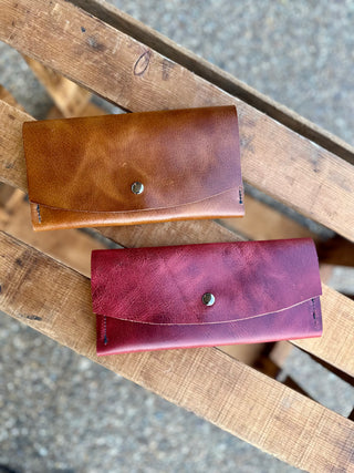 Two handcrafted leather wallets with snap closures displayed on weathered wooden slats outdoors.