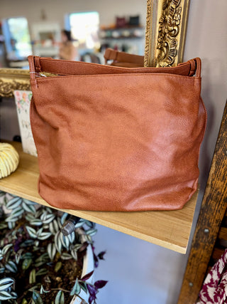 Brown leather tote bag with handles displayed on wooden shelf in retail store setting.