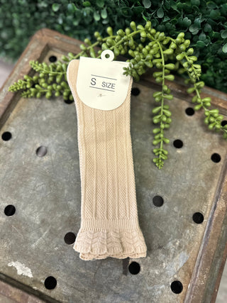 Beige cable knit knee high socks with ruffled cuff displayed on wooden surface with greenery