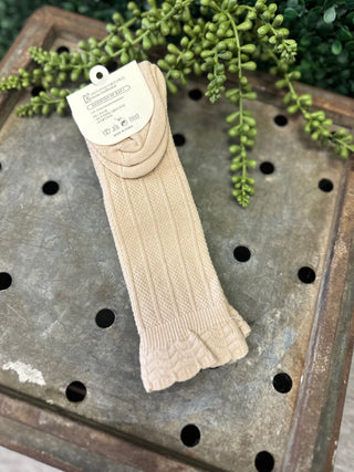 Beige cable knit knee high socks with product tag displayed on rustic wooden surface with plants.