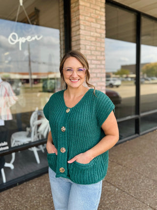 Woman wearing a chunky knit button-down sweater vest with gold buttons and front pockets.