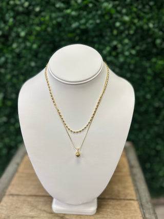 Golden layered necklace set with chain and pendant displayed on white jewelry bust outdoors.