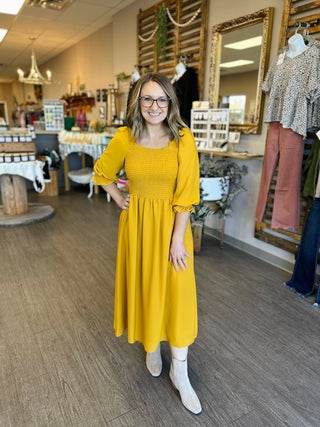 Woman wearing yellow smocked midi dress with puffy sleeves and boots in boutique store setting