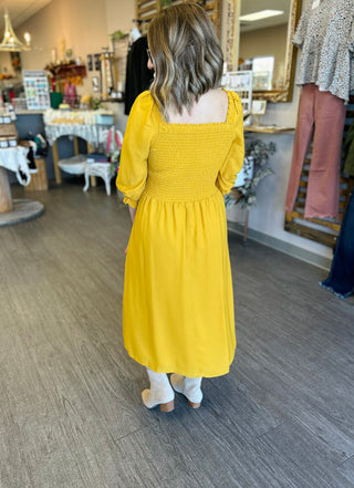 Woman wearing yellow smocked midi dress with long sleeves, shown from behind in boutique setting.