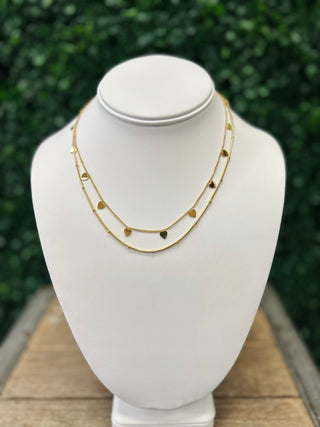 Golden layered necklace with heart charms displayed on white jewelry bust with green foliage background.