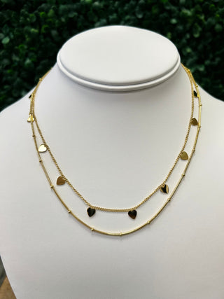 Golden layered necklace with heart-shaped charms displayed on white jewelry bust against green foliage background.