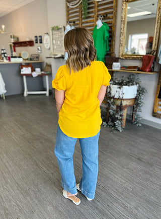 Woman wearing yellow ruffle-sleeve top with jeans, shown from behind in boutique setting.