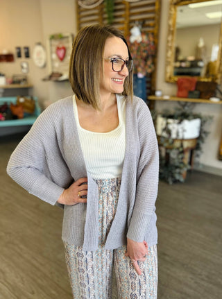 Woman wearing a gray knit cardigan over white top and patterned skirt in casual indoor setting.