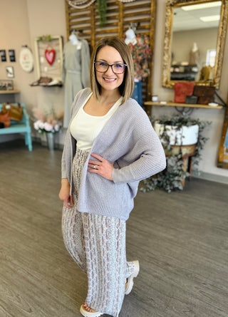 Woman wearing lilac dolman cardigan over white top and floral pants in boutique store setting