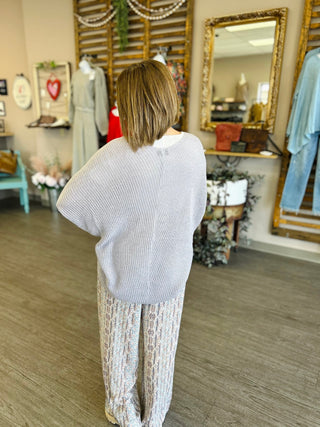 Woman wearing oversized knit dolman cardigan with patterned pants, shown from behind in boutique setting.