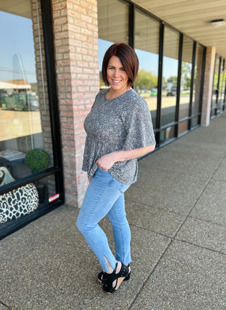 Woman wearing gray speckled babydoll top with jeans, standing on sidewalk outside storefront.