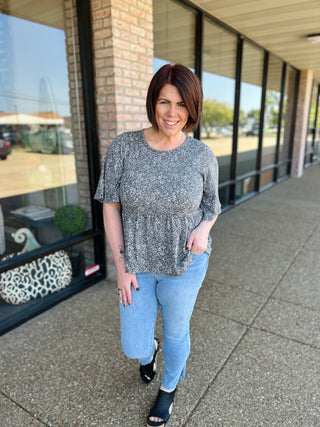 Woman wearing gray speckled babydoll top with jeans, standing on sidewalk outside storefront.