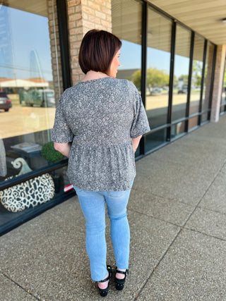 Woman wearing gray floral babydoll top with blue jeans, shown from behind on covered walkway.