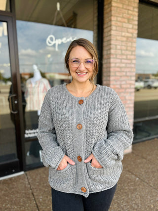 Smiling woman wearing a chunky knit button-front cardigan with front pockets, standing outside a boutique.