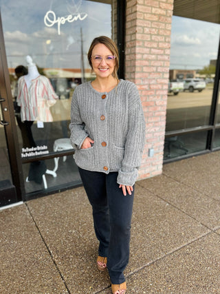 Smiling woman wearing a chunky knit button-front cardigan with pockets, paired with dark jeans.