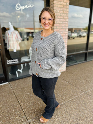 Woman wearing a chunky knit button-front cardigan with wooden buttons and front pockets.