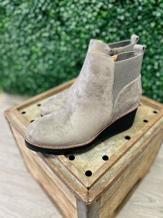 Grey faux suede ankle booties with elastic side panels displayed on rustic wooden crate with green foliage background.