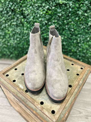 Grey suede ankle boots with side zippers displayed on wooden tray against green backdrop