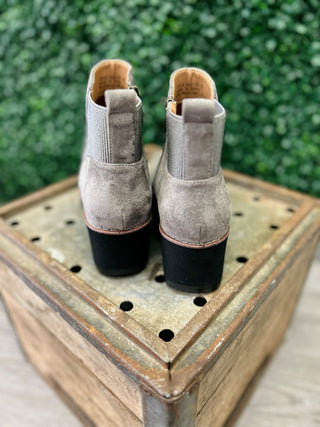 Grey suede ankle booties with block heels displayed on wooden crate against green foliage background.